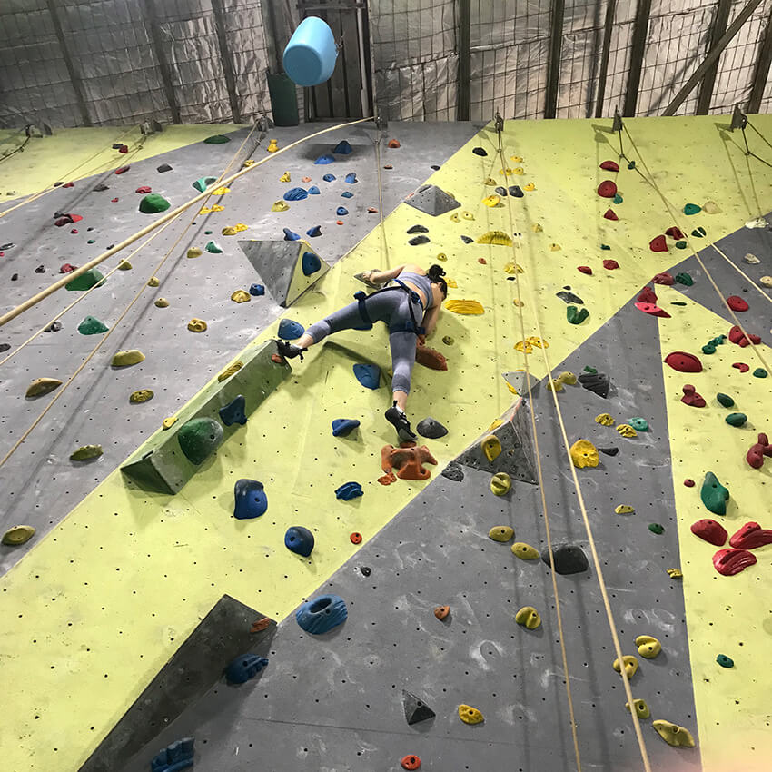 A woman wearing blue leggings and a sports bra climbing an indoor rock climbing wall, facing the wall. She has her feet on orange holds and there are many different coloured holds on the wall, which is grey and yellow.