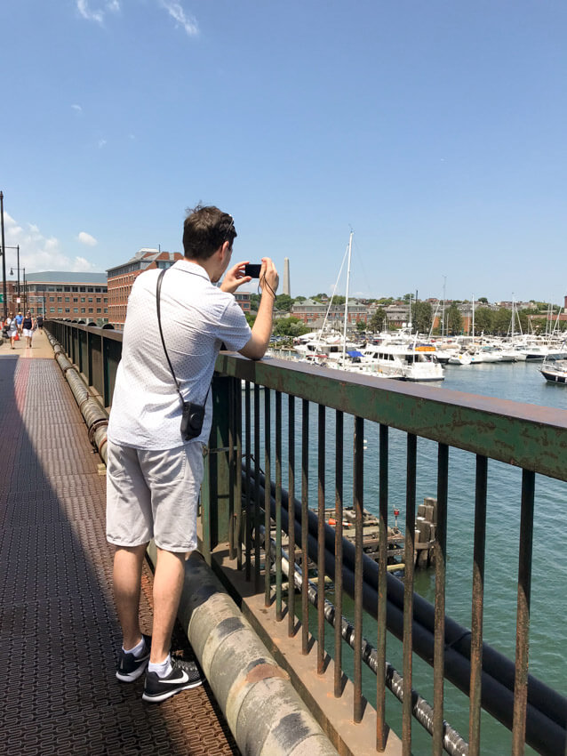 A man in shorts and a white shirt, taking a photo on a camera over the railing of a bridge. He is photographing some boats in a harbour