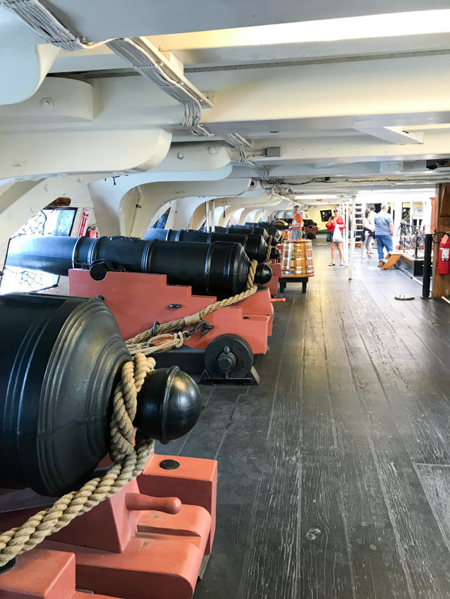 The interior of a ship, with low ceilings. There are black cannons down the left hand side