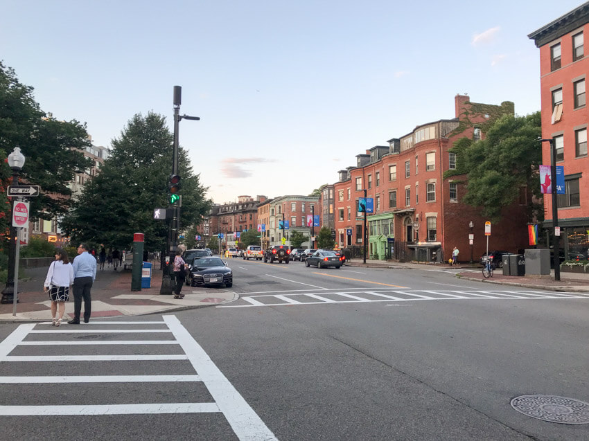 An early evening photo taken from the intersection of a street. Two crosswalks can be seen, and in the street ahead some cars can be seen as well as some historic-looking brick buildings