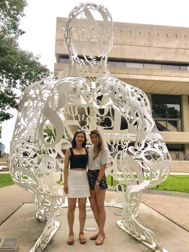 Two women, smiling, wearing summer clothing and sandals with an arm around each other. They are standing in front of a large sculpture resembling a person’s head and hands, made out of white-painted steel numbers and letters