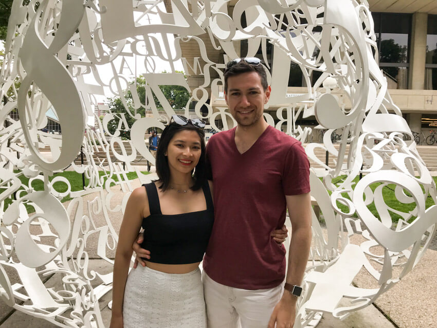 A woman and man smiling, with an arm around each other, in front of a sculpture made out of white-painted steel letters and numbers melded together