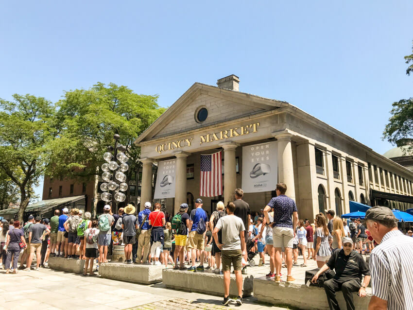A long building with a pillared entrance. Above the pillars are the words “Quincy Market”. There are many people gathered around the front of the building.