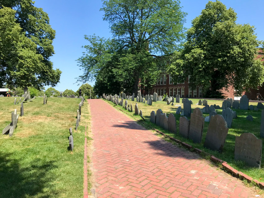 A red brick path through a cemetery, where headstones sit in lines on green grass
