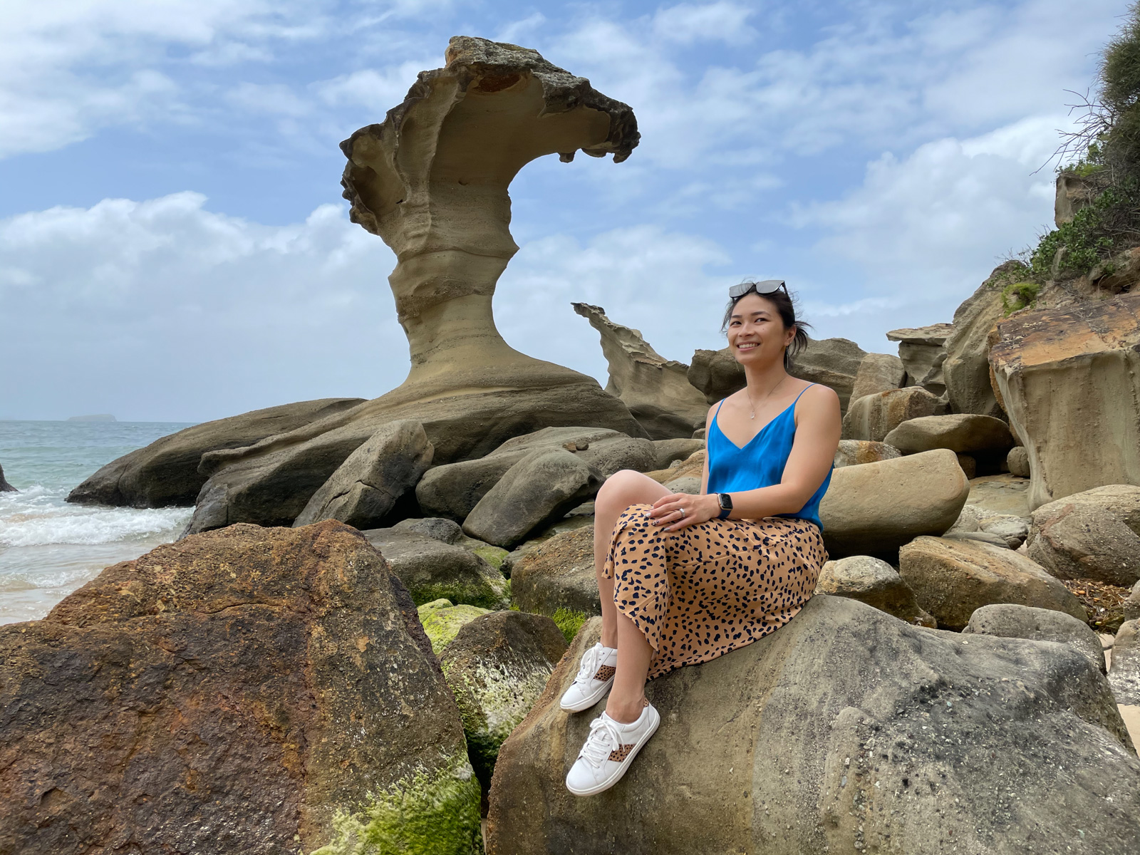 A woman with dark hair, wearing a blue top and a long cheetah print skirt with white sneakers, sitting on a rock by the beach. Behind her is a giant rock formation like a wave. The sky is cloudy