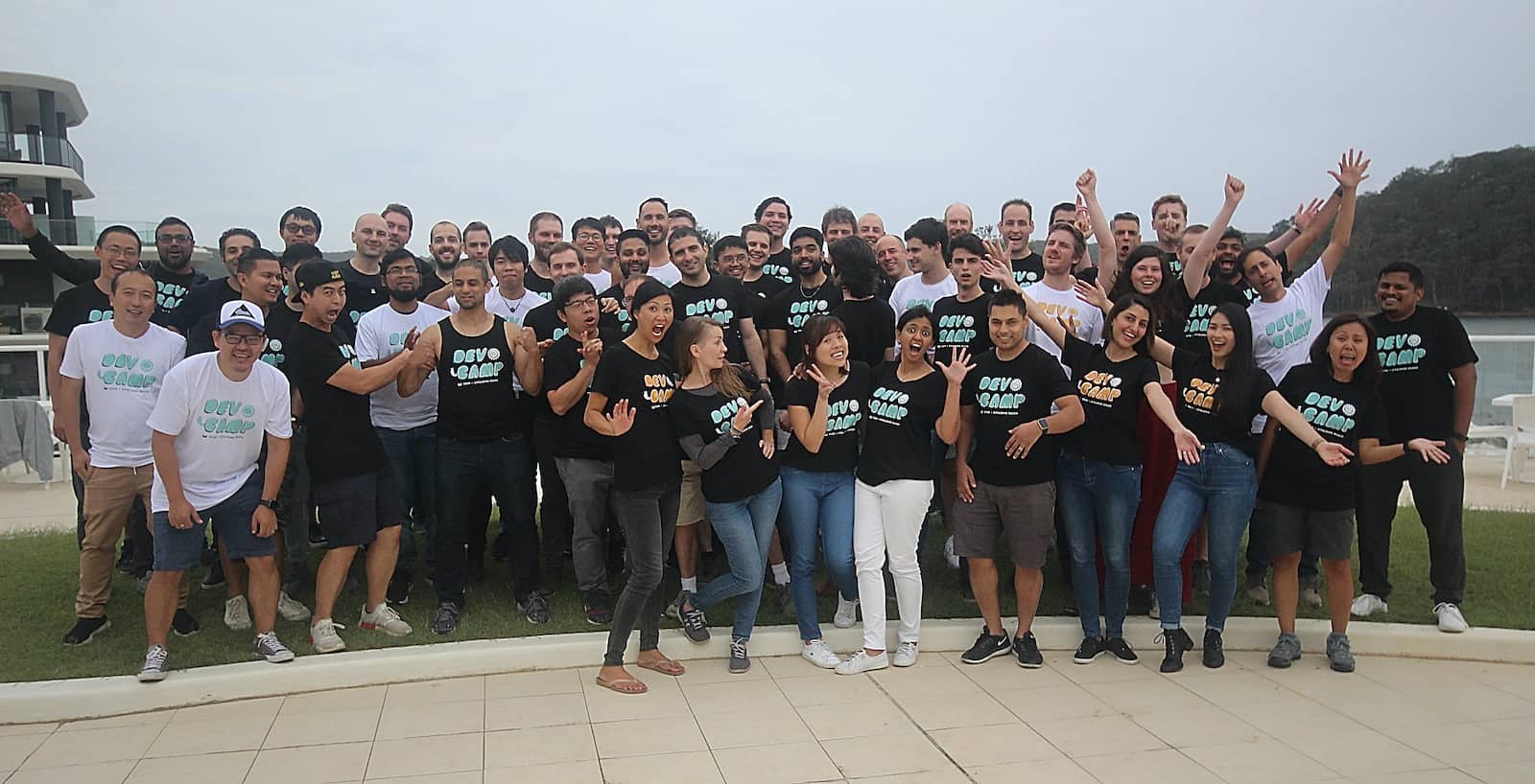 A group of people wearing shirts with matching bubble text reading “Devcamp”, some people with their hands in the air. The sea can be seen behind them
