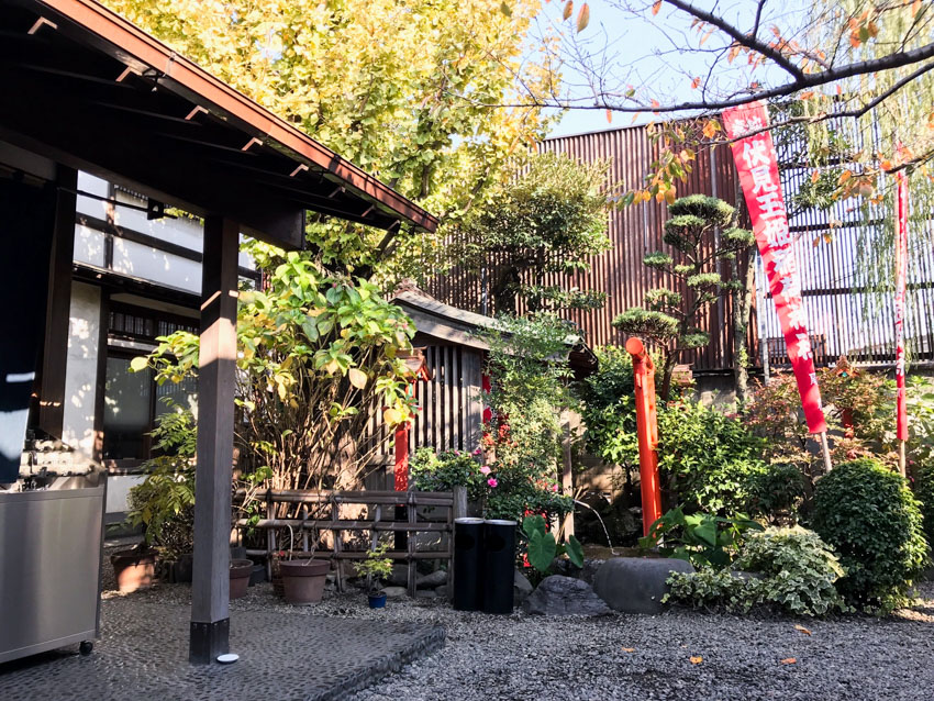 A garden area with small grey stones lining the ground, tall and thin red flags with white Japanese characters on them, plenty of small green plants and trees, some concealing a small hut. In the foreground is the awning of a building.