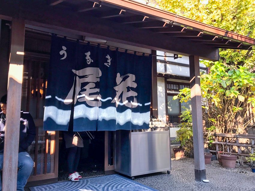 The front of a traditional Japanese resturant, with a short curtain with slits, partially cover ing the entrance. The curtain is navy blue in colour and has Japanese kanji characters on it in white. Someone is standing behind the curtain and their legs and feet can be seen. There is a wooden awning and some green plants in the background.