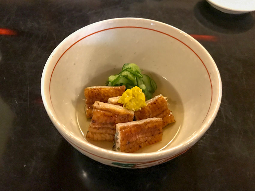 A small cream-coloured bowl filled with pale yellow soup, and several slices of grilled eel that are a golden brown in colour. There is some yellow garnish on top and some green sliced cucumber.