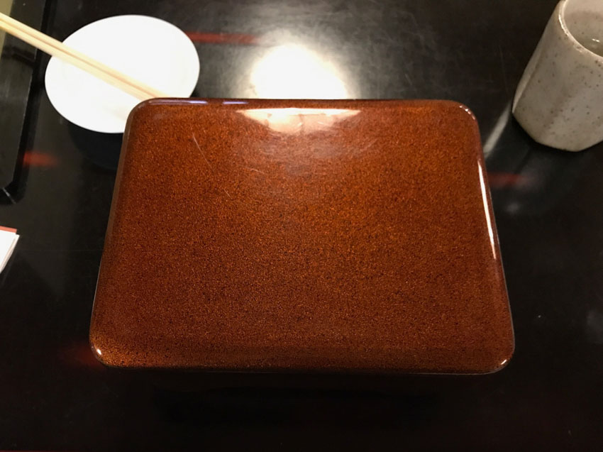 A rectangular box with rounded corners, sitting on a table. The box has a slightly shiny surface. In the background is a grey cup filled with water, and a pair of chopsticks.