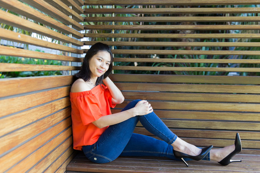 A girl sitting in a wooden cabana sideways with one leg outstretched and one knee bent, smiling. She is wearing an orange top, dark blue jeggings, and black high heels. Her left hand is held behind her head and the other hand on her raised knee.