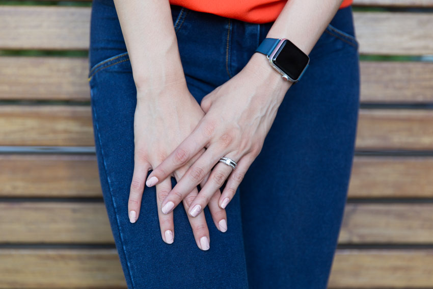A girl’s left hand on top of her right hand, resting on her thigh as she is standing. She is wearing silver diamond rings on her left ring finger and a smartwatch with a blue band.