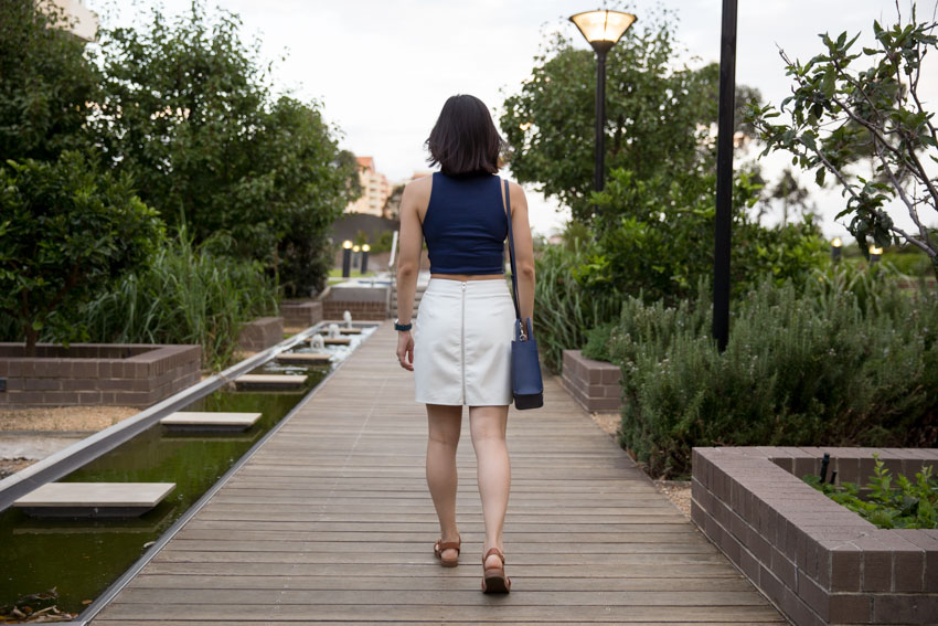 A girl walking on a path of wooden slats, wearing a white skirt that zips all the way up the back. She has short dark hair and is carrying a small navy handbag off her shoulder.