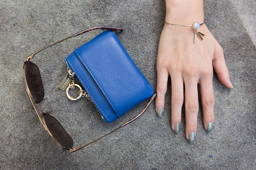 Flatlay style of my sunglasses and blue wallet with my hand in view, with my painted nails