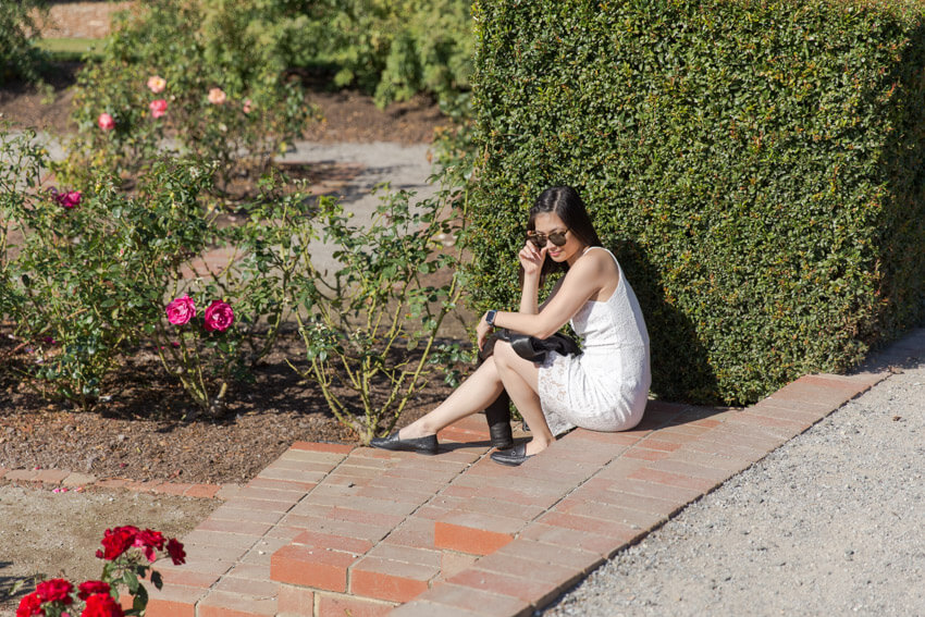 A woman with short dark hair and sunglasses, in a sleeveless white dress sitting on a step in a rose garden. She has a black jacket on her lap and is holding the frame of her glasses with one hand.