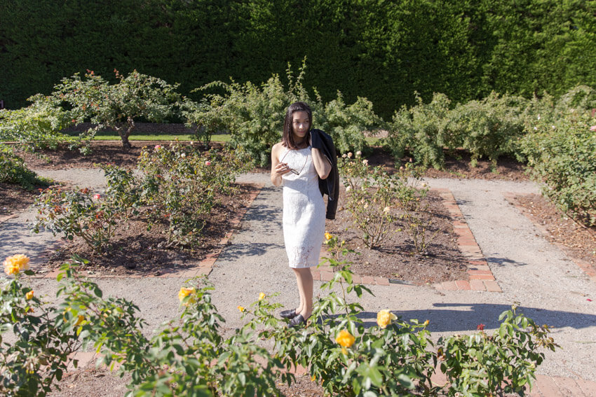A woman in a strapless white dress standing in the dirt paths of a rose garden. There are small garden inlays in the dirt paths. The woman is holding her sunglasses between her fingers and with the other hand has a leather jacket slung over her shoulder