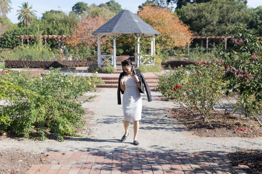 A woman in a white dress with a black jacket draped over her shoulders, looking down, mid-step. She is wearing black loafers and in the background is a gazebo and a rose garden.