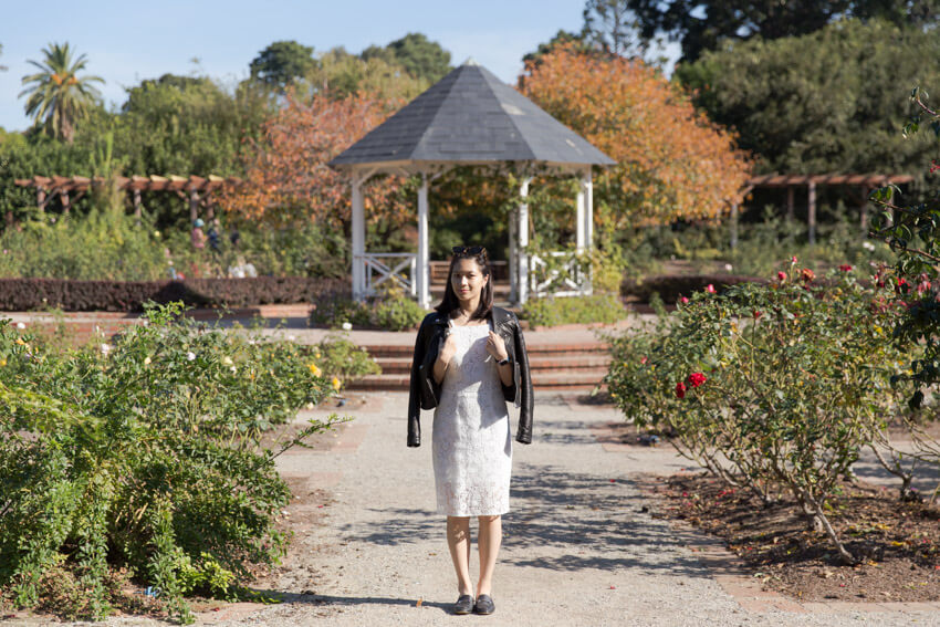 A woman in a white dress with a black jacket over her shoulders, facing the camera. In the background is a gazebo and a rose garden.