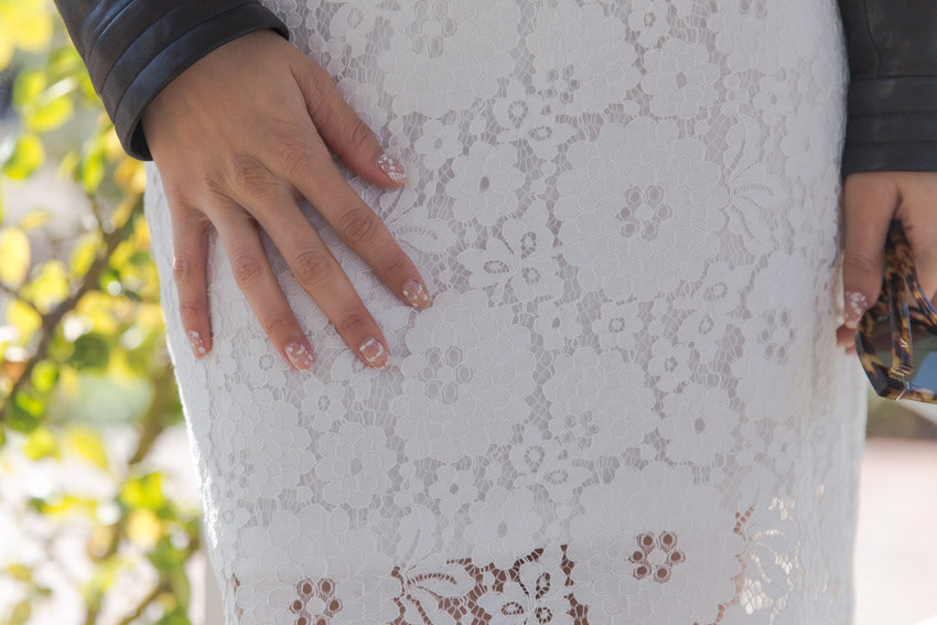 A close, front-on shot of a woman’s hand against the white lace dress she is wearing. Her fingnernails have a white floral design that still shows the bare nail where the design is not printed.