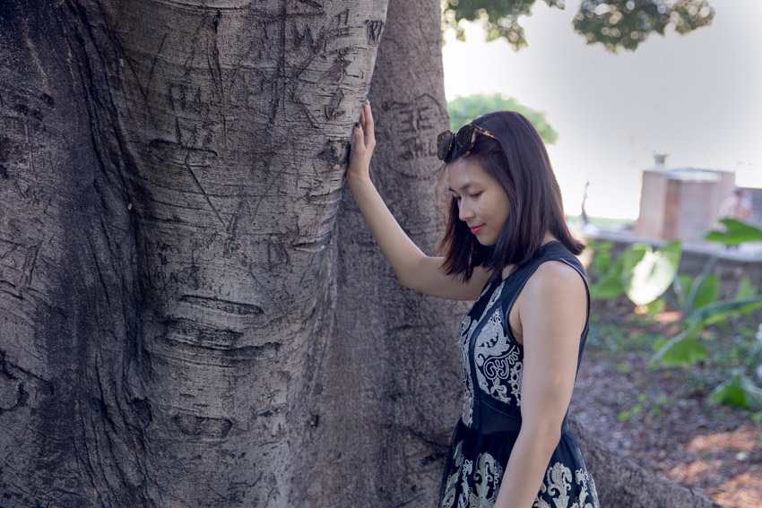 A woman with sunglasses on top of her head, standing by a large tree trunk. She has her right hand resting on the tree. She is wearing a sleeveless black dress with gold embroidery.