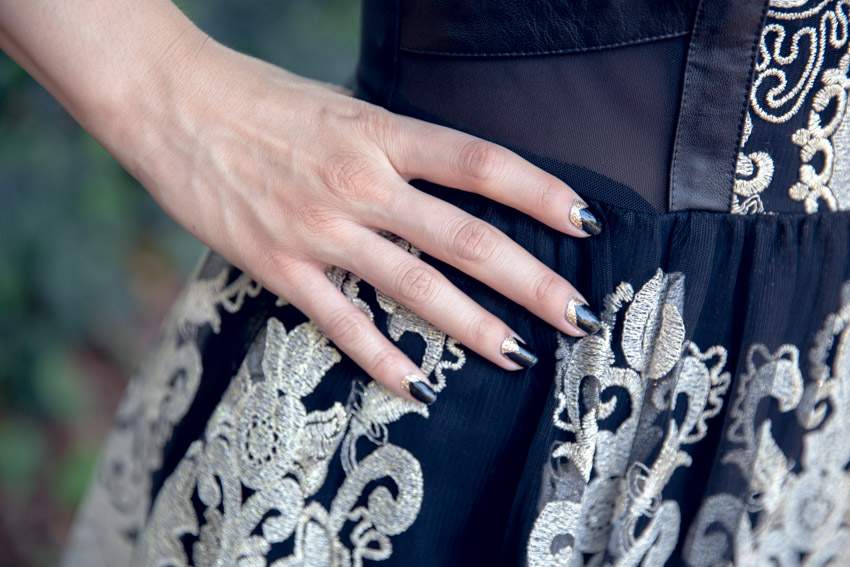 A close-up of a woman’s right hand on her hip. She has black nails with gold glitter chevron accents. The dress she is wearing is made of black mesh and gold embroidery.