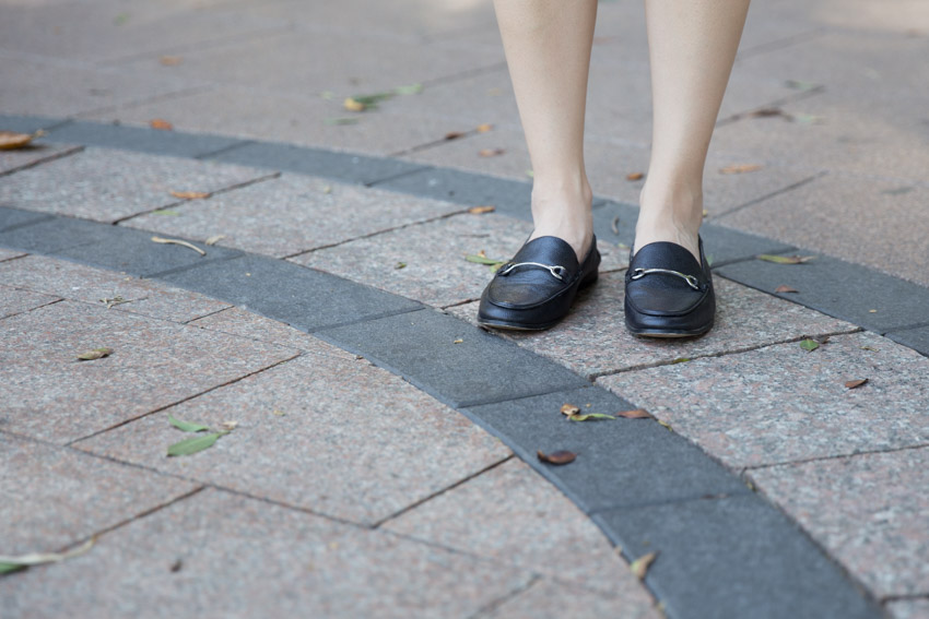 A woman’s feet with black loafers, standing on black and light brown tiled pavement with the tiles laid out in concentric circles. Some leaves sit on the pavement.
