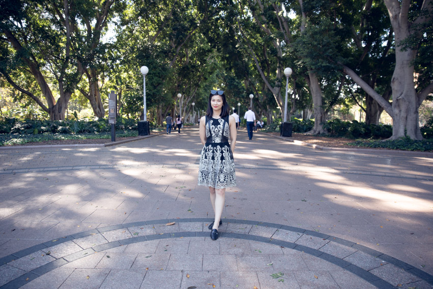 A woman wearing a black and gold sleeveless, knee-length dress. She has short dark hair and is wearing sunglasses on top of her head. She has her hands behind her back and is wearing black loafers. She is standing at the entrance to a park with a wide path extending behind her, lampposts on the edges of the wide paths and many tall trees in the background