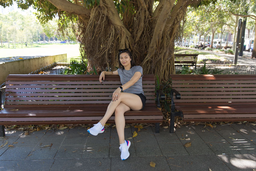 Me sitting on a brown wooden bench, with my left leg crossed over my right. My left hand rests on my left knee and my right arm is draped over the back of the bench. I am looking somewhere beyond the camera.