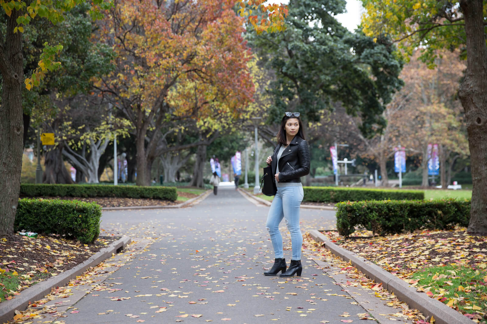 A woman in light blue jeans and a black leather jacket, holding a handbag over her right shoulder. Her left side faces the camera. She is standing on a path surrounded by trees with yellow and green leaves.