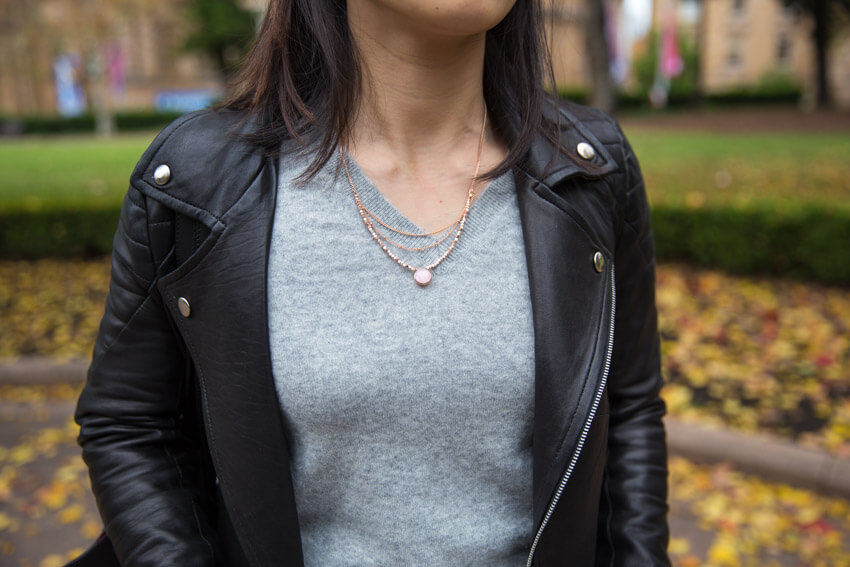 A close shot of a woman in a grey v-neck sweater wearing a rose gold necklace with layered chains and small beads, and a pink-coloured pendant drop