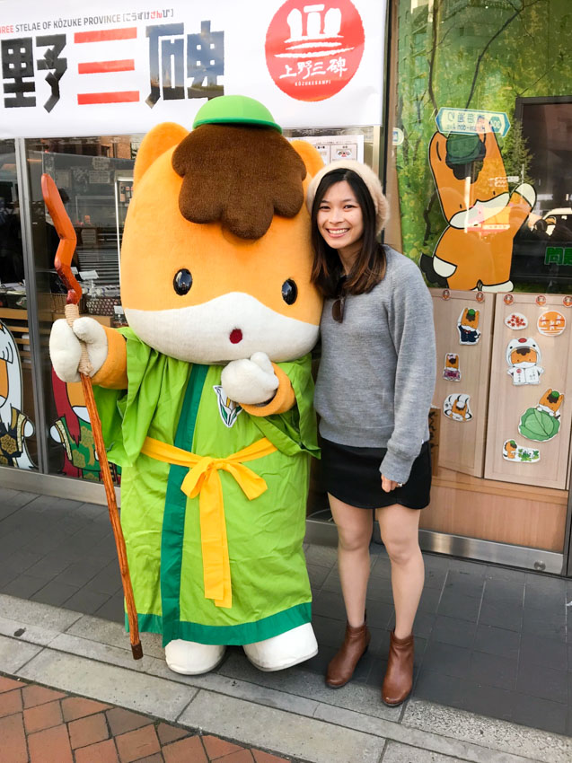 A girl in a grey jumper, black skirt and brown boots, smiling, alongside someone dressed in an animal costume. The animal is a bit like a cat with orange fur and white paws, and is wearing a bright green toga and small bright green cap.
