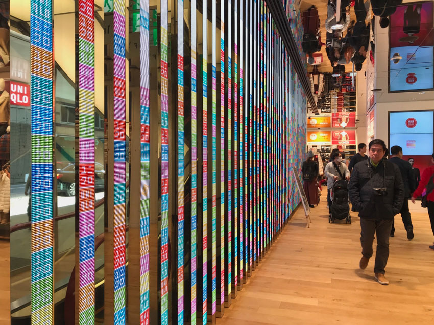 The inside of a store with wooden floors. Narrow reflective panels alternated with panels of columns of coloured squares make up a wall on the left. Some people are visible in the distance, not too close to the wall.