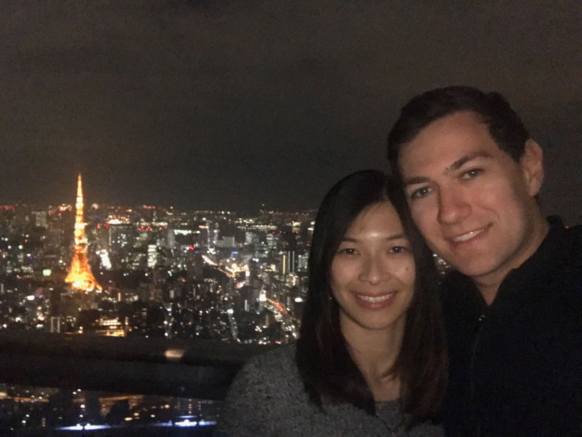 A girl with short dark hair, and boy with dark hair, smiling. It’s nighttime and the lights are on in the buildings that are in the background. There is a tower that stands out with yellow/orange lights.