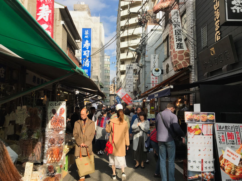 A market during the day, filled with people. Dried octopus is hanging from a stall on the left, and a stand outside with a menu is visible on the right. Lots of power lines can be seen above the stalls and some apartment buildings in the distance.