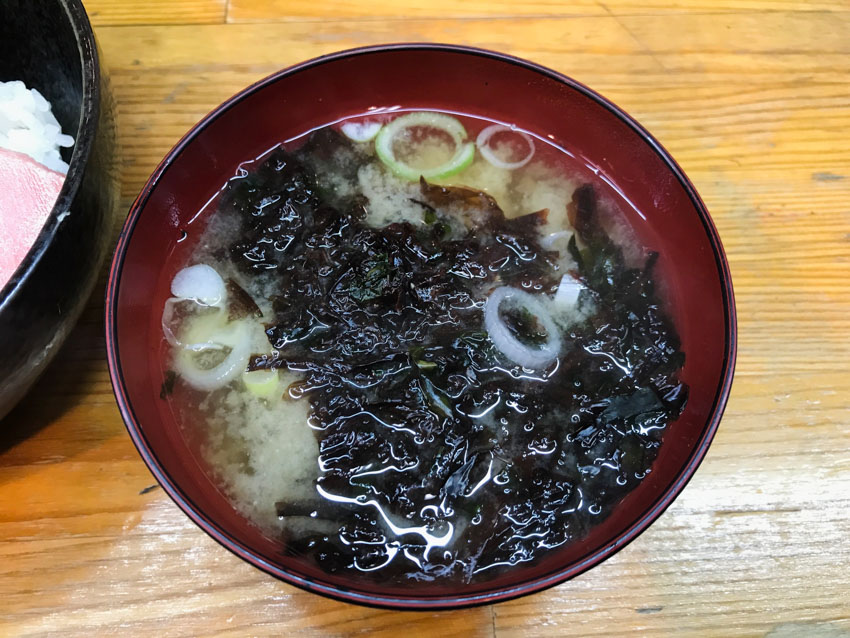 A bowl of cloudly soup with small green onions. There is a lot of dark green wet seaweed in the bowl.