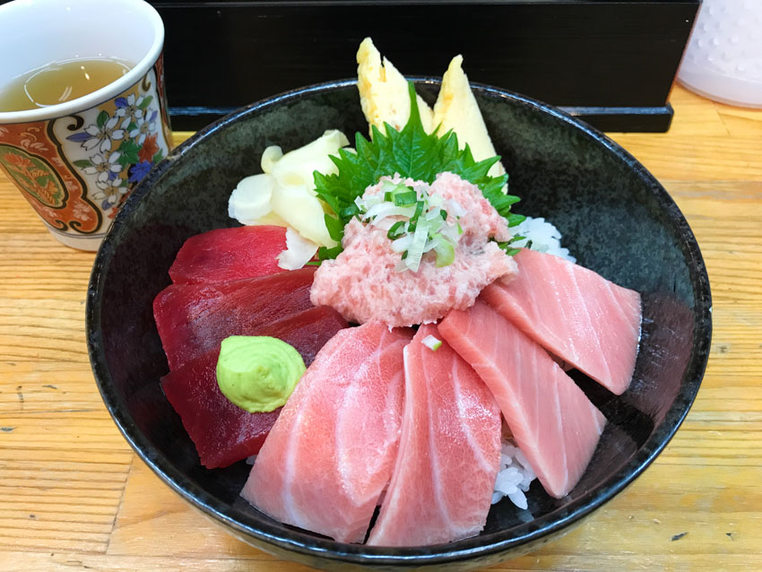 A black bowl containing rice with deep red and light pink slices of raw tuna. It’s topped with a soft scoop of pink tuna, yellow pickled ginger, and a pea-sized piece of wasabi.
