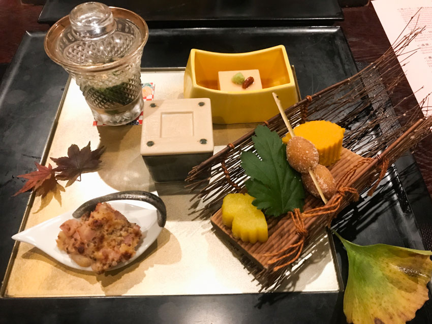 A tray of appetisers, one in a small glass with a lid, another in a square crucible with a lid, a square of tofu in a small rectangular cup, a leaf-shaped dish with an appetiser of shrimp with bacon and cheese, a small sled made of natural sticks with a couple of cakes and a skewer
