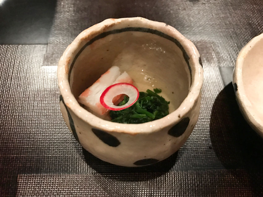 A deep small stone bowl with a piece of crab, greens, and a slice of radish