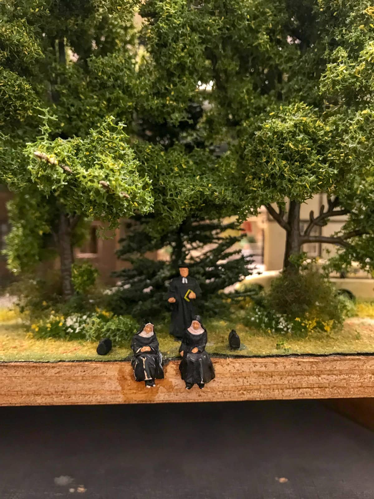Two figurines of nuns sitting at the edge of a landscape. Behind them is a figurine man dressed in black. There are trees behind them as well