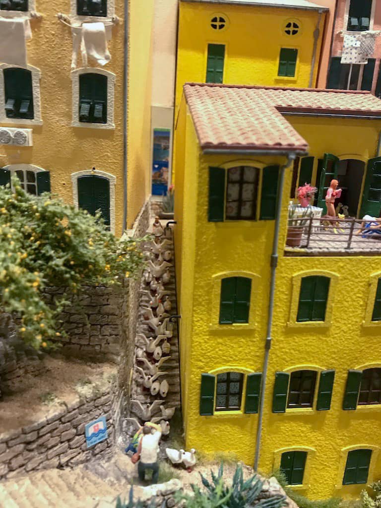 Old Italian-style miniature houses with a long flight of stairs down the side. On the flight of stairs are many miniature seagulls/ducks with a figurine man taking a photo from the bottom of teh stairs.