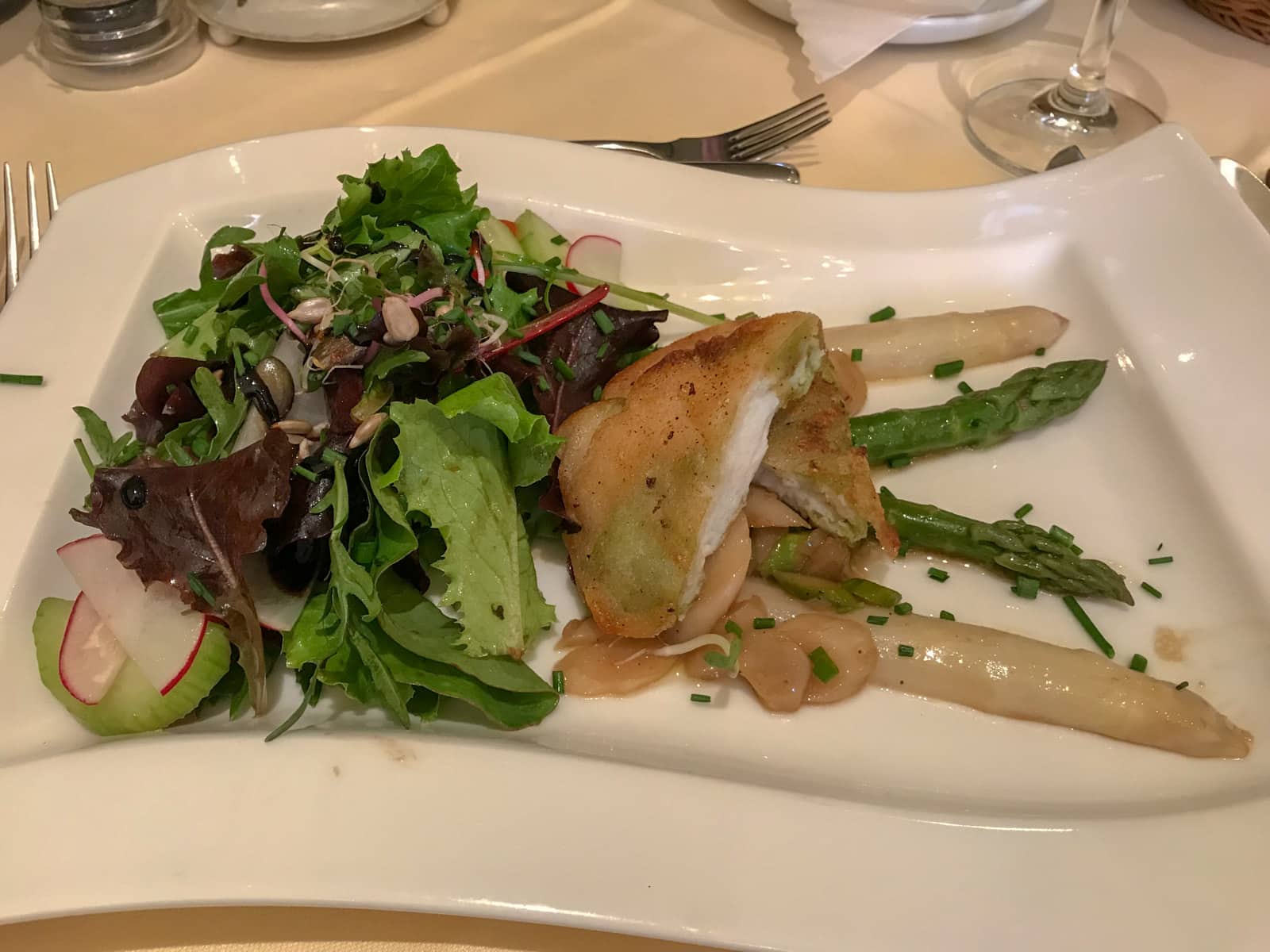 A white, flag-shaped plate on a dining table with salad, asparagus and fish served on it.