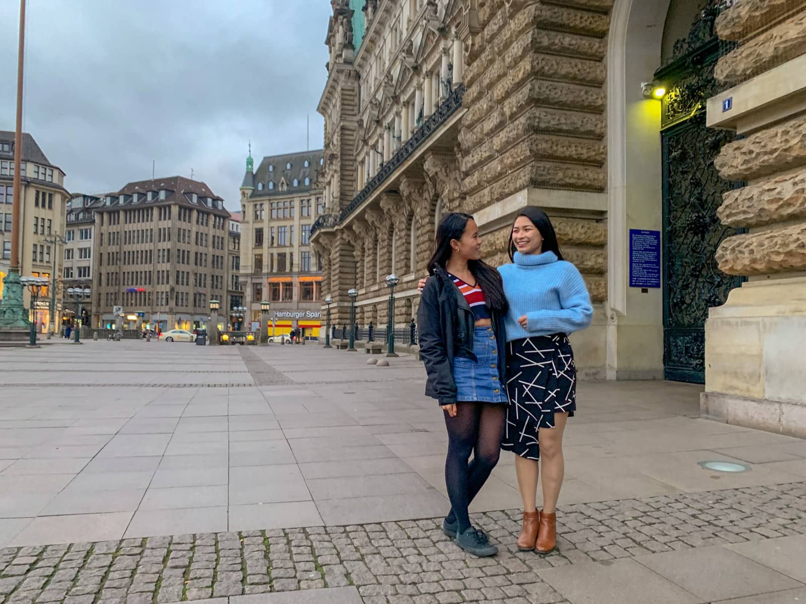 Two women smiling, with their arms around each other. Both have dark hair. One is wearing a striped coloured top and a black jacket with a blue skirt, the other is wearing a light blue sweater with a black-and-white patterned skirt. They are in a street setting in Hamburg, Germany