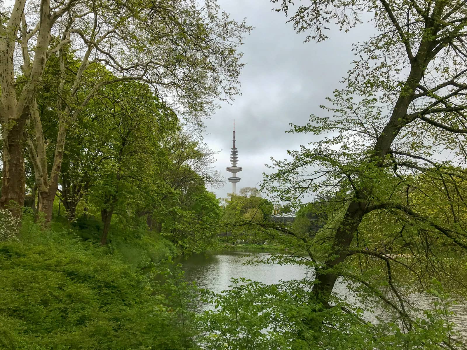 The Hamburg Hertz Tower in Hamburg, Germany. It’s a telecommunications tower as seen from between trees inside of a park. It’s a very cloudy day.