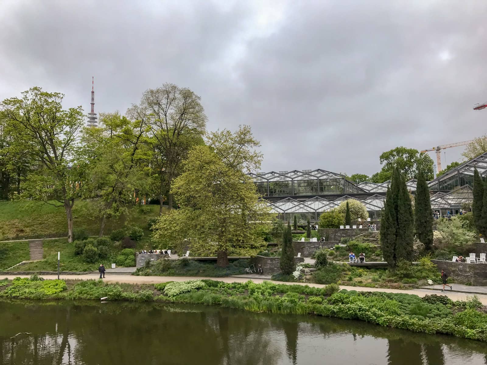 The inside of a park, with a river in the foreground, and greenhouses in the background. There are many trees in the park