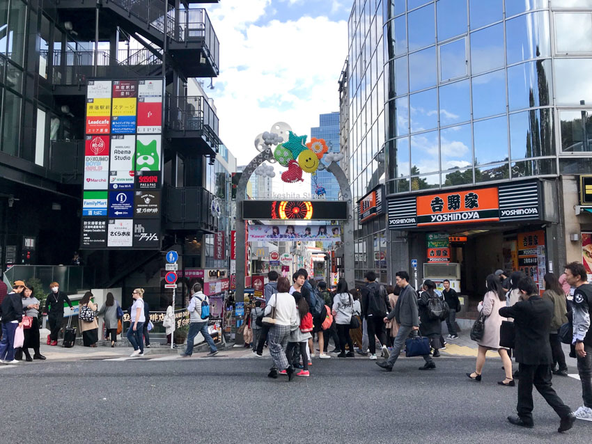 The entrance arch to Takeshita Street, with many pedestrians walking. On the left, there is a building with a metal staircase, and stairs leading downwards, and on the right, a glass high-rise building and a Yoshinoya restaurant