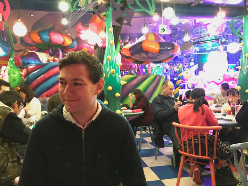 Nick looking off-camera, in a restaurant setting with a black-and-white checkered floor and colourful decor, some resembling big trees and others looking like toadstools