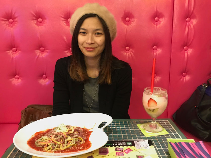 Me sitting at a table, with a white palette-shaped dish of colourful spaghetti pasta in red saurce in front of me. A drink in a round wine-shaped glass also sits on the table.