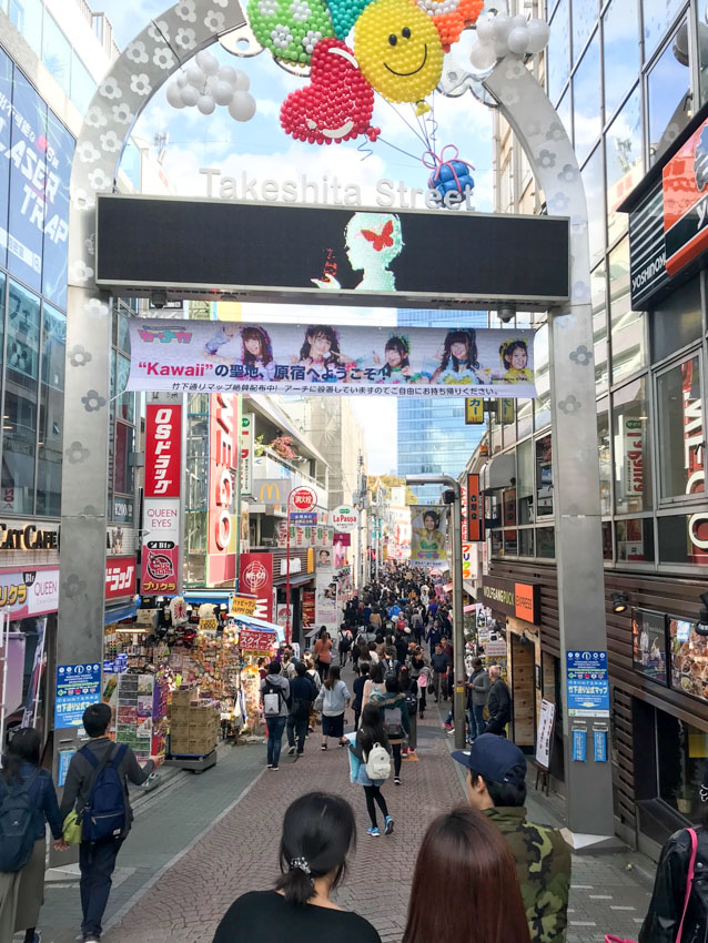 The entrance arch to a busy street, called Takeshita Street. The arch has a banner hanging towards the top, an LED screen with advertisements, and colourful balloons creating a smiley shape and heart shape at the top of the arch