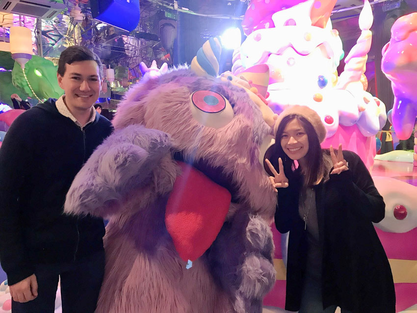 Me and Nick, smiling, standing on either side of someone dressed in a giant fluffy light purple monster costume. The monster has a large tongue and big eyes. I am making two peace signs with my hands.