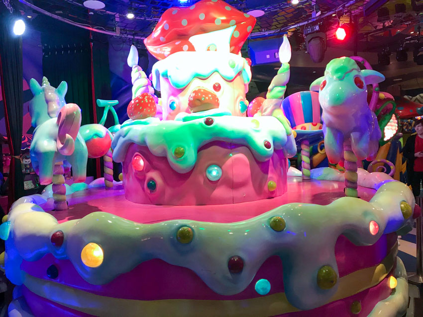 A sculpture made from what appears to be shiny plastic, resembling a cake with thick icing and horses like a carousel. Round coloured lights on the sculpture and hanging above the sculpture light it up